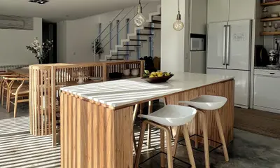 Cocina integrada con isla de madera, banquetas blancas y una escalera al fondo.